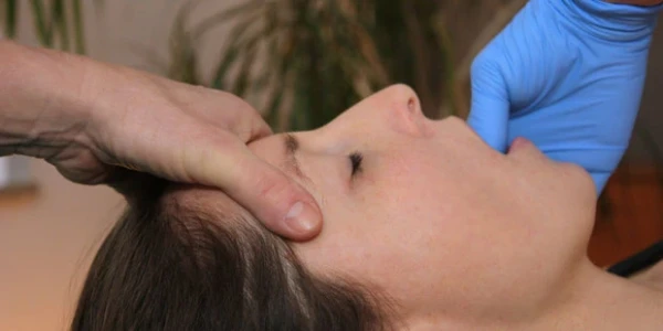 Eine seitliche Aufnahme zeigt eine liegende Person mit geschlossenem Auge. Eine Hand stabilisiert den Kopf, während eine zweite Hand in blauen Einmalhandschuhen in den geöffneten Mund greift, um therapeutischen auf den Kiefer einzuwirken.