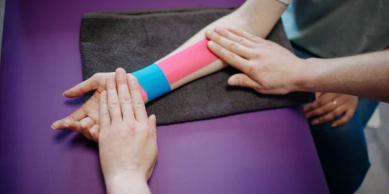 Eine Nahaufnahme von einer Person, die Physiotherapie erhält, mit leuchtend pinkem und blauem Kinesiologie-Tape am Unterarm, während eine andere Person sanft das Handgelenk und die Hand stützt.
